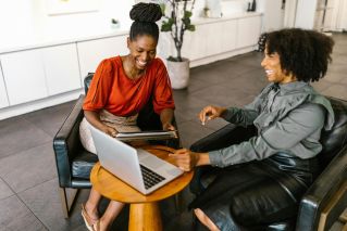 Two businesswomen in a modern office environment discussing work over a laptop.